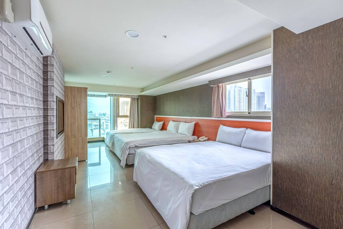 A hotel room with two beds, a TV, a wooden side table, and a large window with a city view.