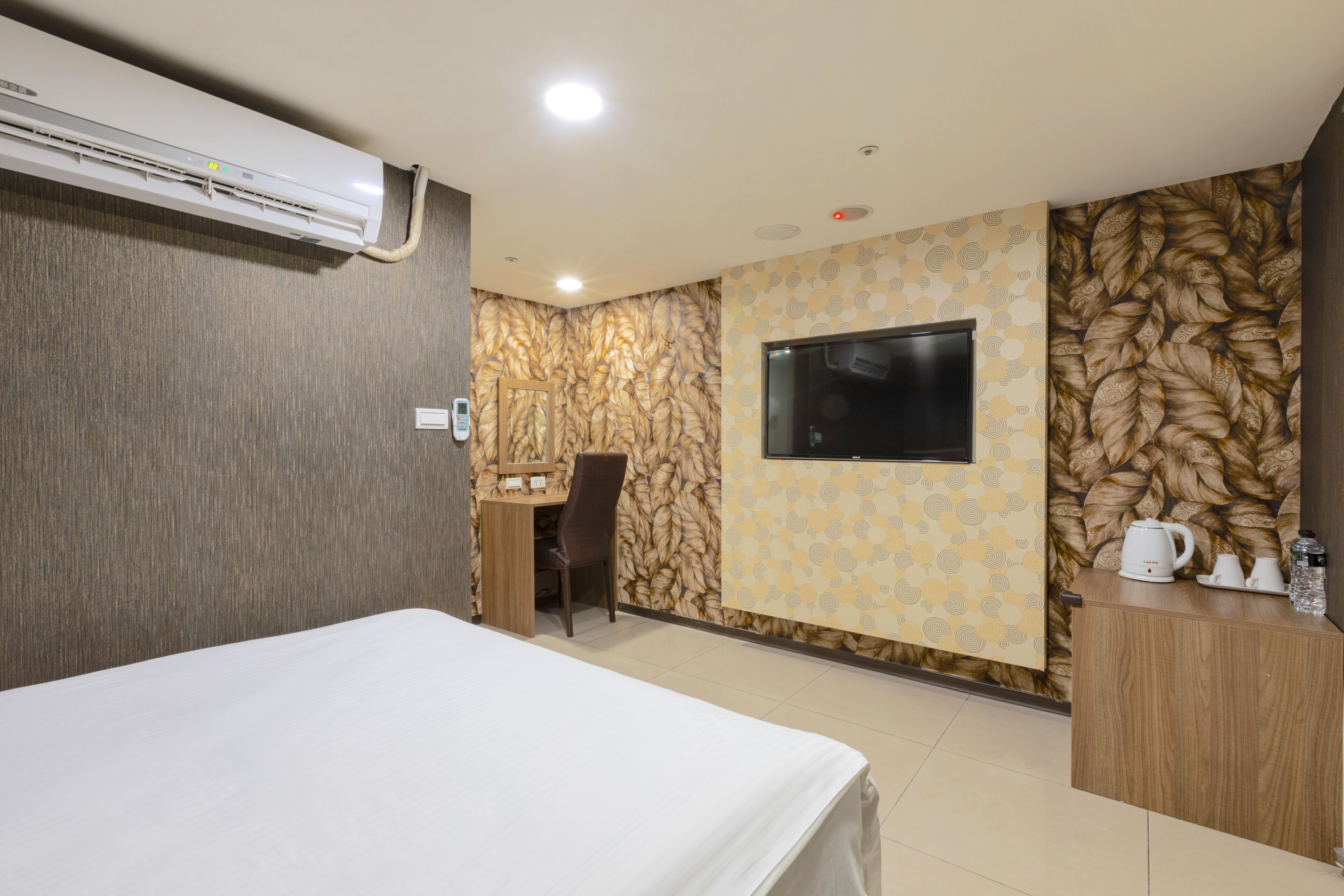 A hotel room with a bed, a desk, a chair, a TV, and a kitchenette area.