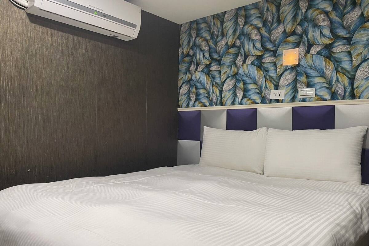 A hotel room with a bed, a wall-mounted air conditioner, and a patterned wallpaper.