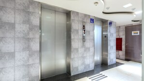Et moderne elevatormiljø med grå fliserede vægge, et digitalt elevatorkontrolpanel og en rød nødudgangsdør.