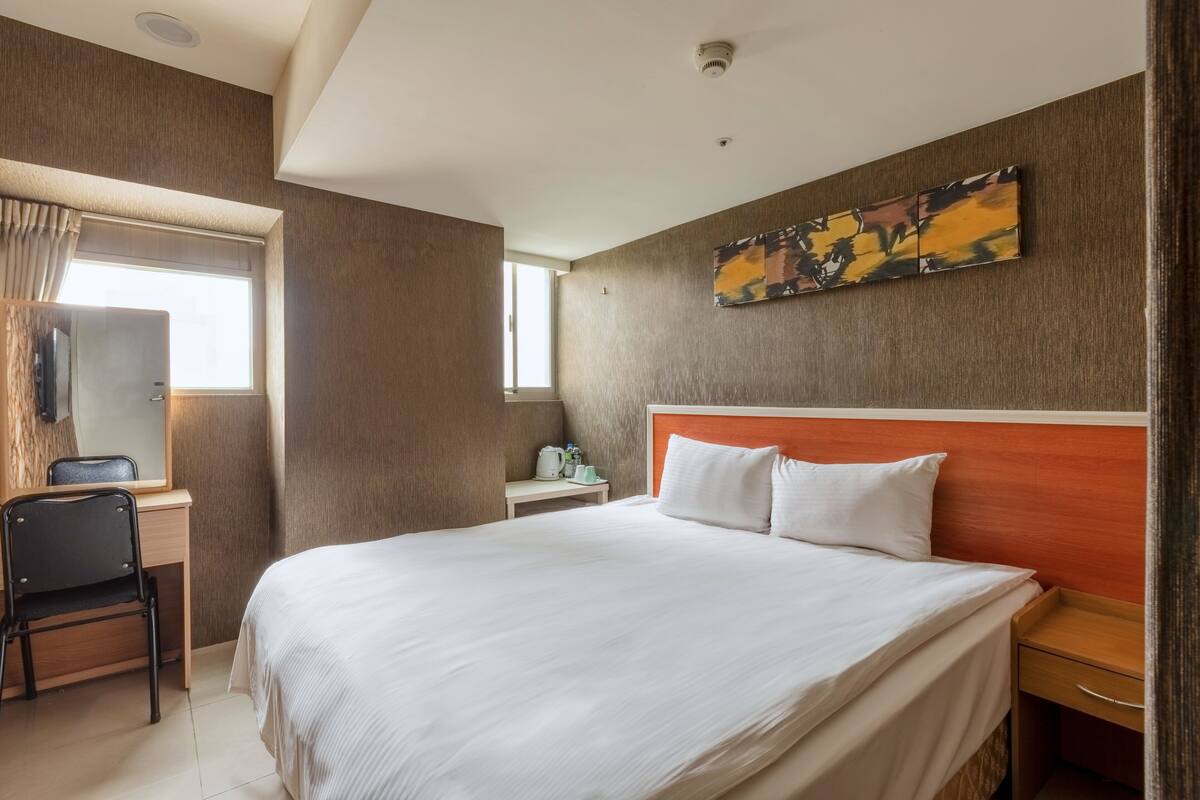 A hotel room with a large bed, a desk with a chair, a small table with a kettle, and a painting on the wall.