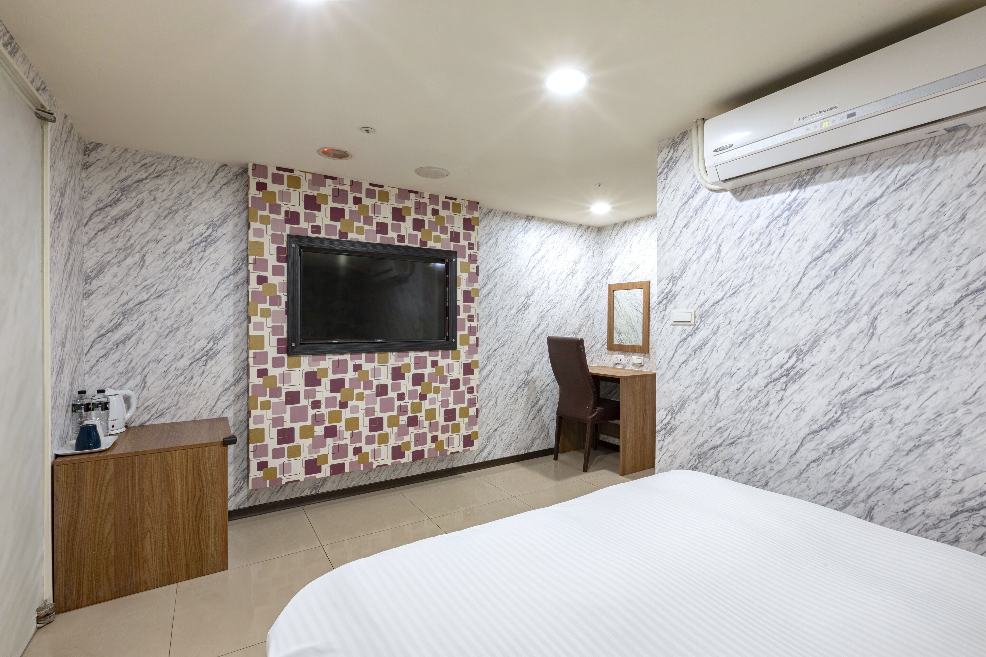 Habitación de hotel con cama, escritorio, silla, televisor montado en una pared de mosaicos y unidad de aire acondicionado.
