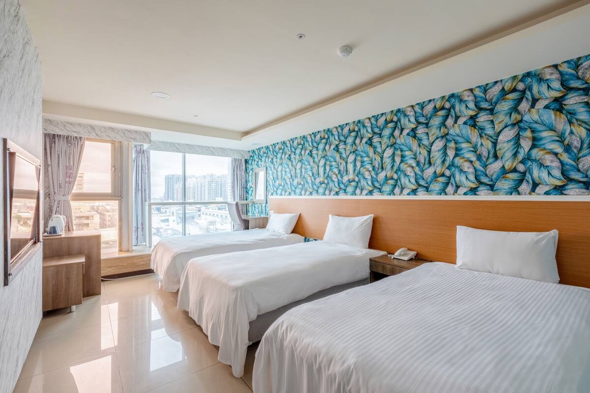 A hotel room with three beds, a large window with curtains, a city view, and a patterned wall.