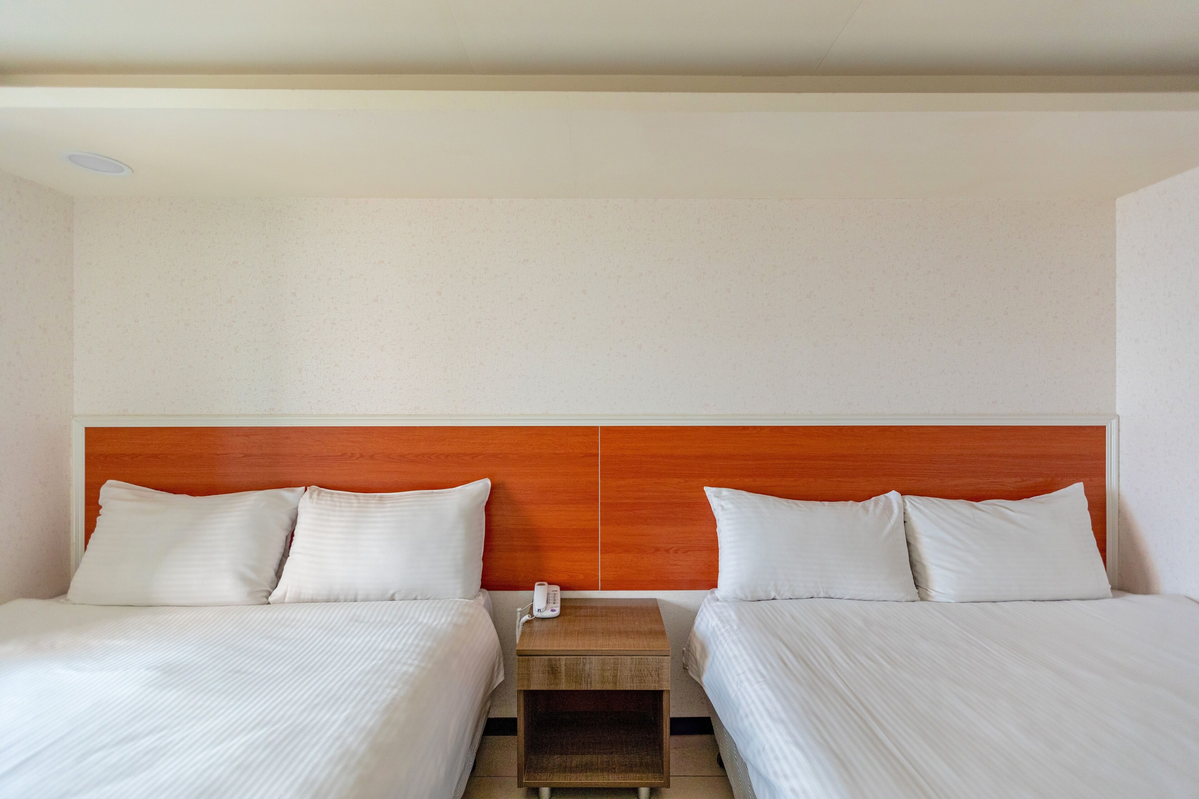 A hotel room with two beds, a wooden headboard, a bedside table with a phone, and a neutral-colored wall.