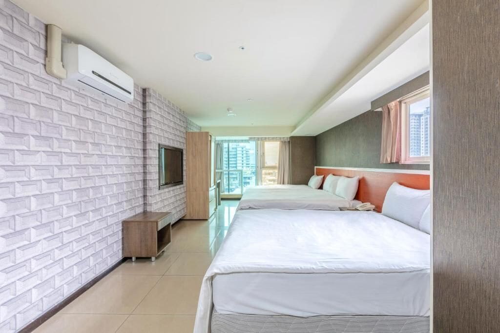 A hotel room with a brick-patterned wall, a flat-screen TV, a bed with white bedding, and a window with curtains.