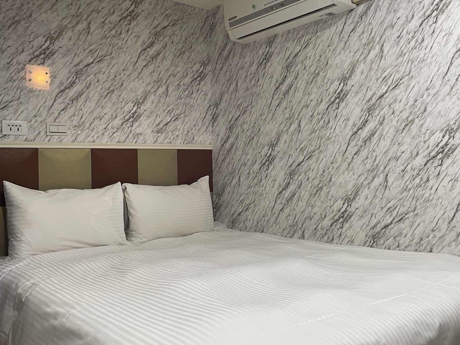 A hotel room with a marble-patterned wall, a bed with white bedding, and an air conditioning unit above the bed.