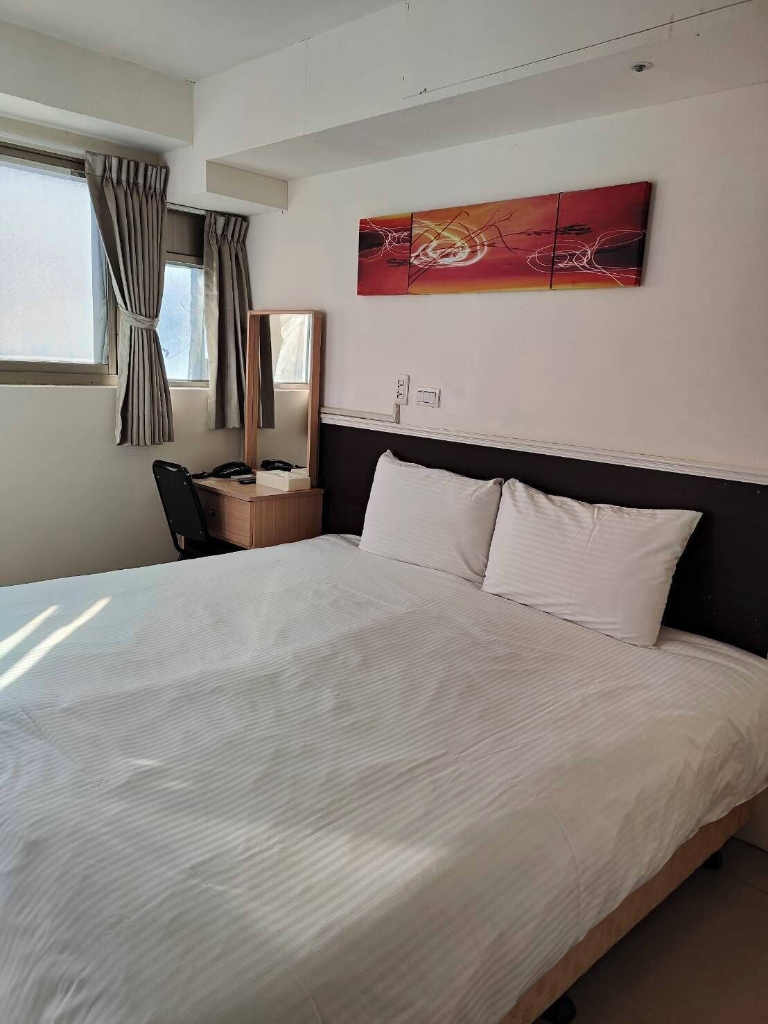 Quarto de hotel com uma cama grande, uma escrivaninha com cadeira, um espelho e uma janela com cortinas.