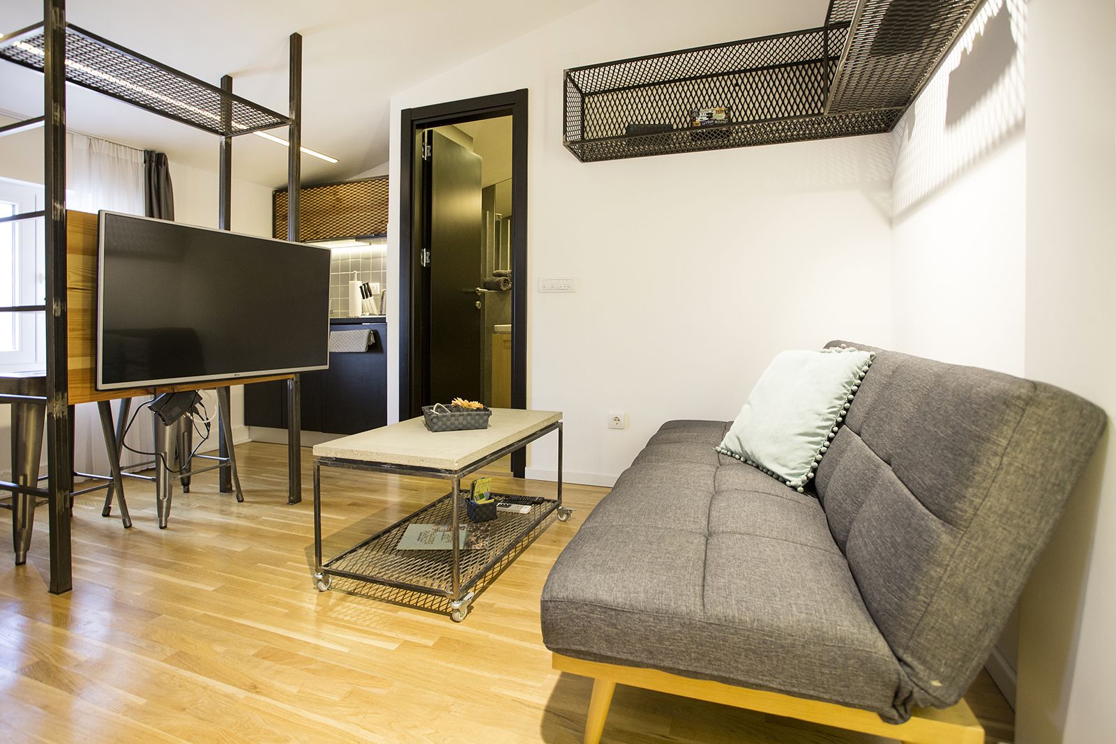 Una sala de estar moderna con un sofá gris, un televisor sobre un mueble bajo, una mesita y una cocina al fondo.