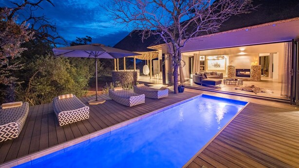 A modern outdoor pool area with wooden decking, lounge chairs, and a well-lit interior visible through an open door.