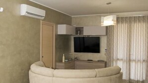 A modern living room with a round beige sofa, a flat-screen TV mounted on the wall, a dining table with chairs, and an air conditioning unit on the wall. - Casa Vacanze Fontana offered from 22/07/1929/07 / 19e from 08/25/19 to 31/08/19  (San Vito Lo Capo)