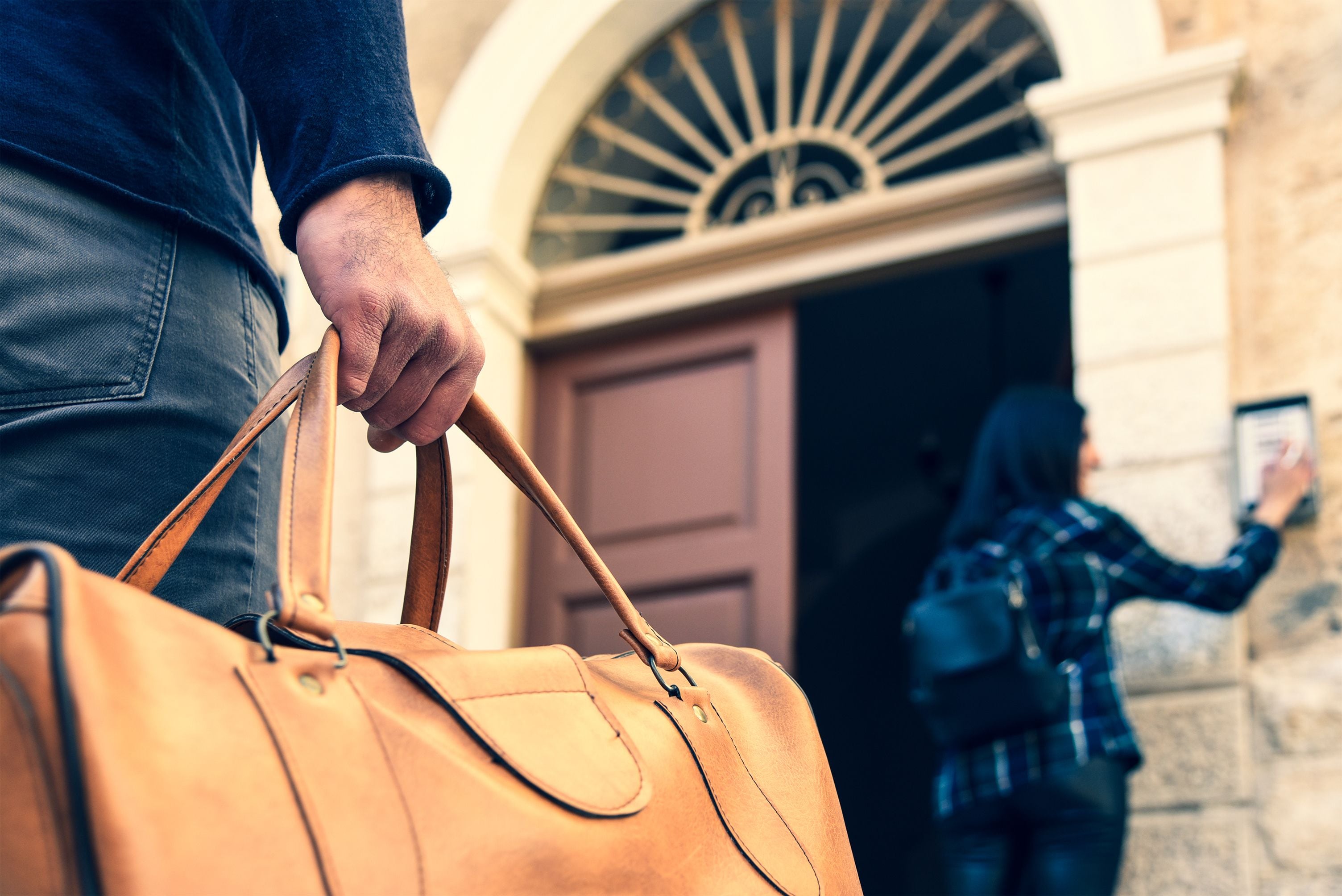 Une personne tenant un sac en cuir marron devant un bâtiment à la façade en pierre et à l’entrée en arc de cercle.