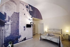 Quarto com cama, mesa de cabeceira, televisão na parede, uma porta e um mural.