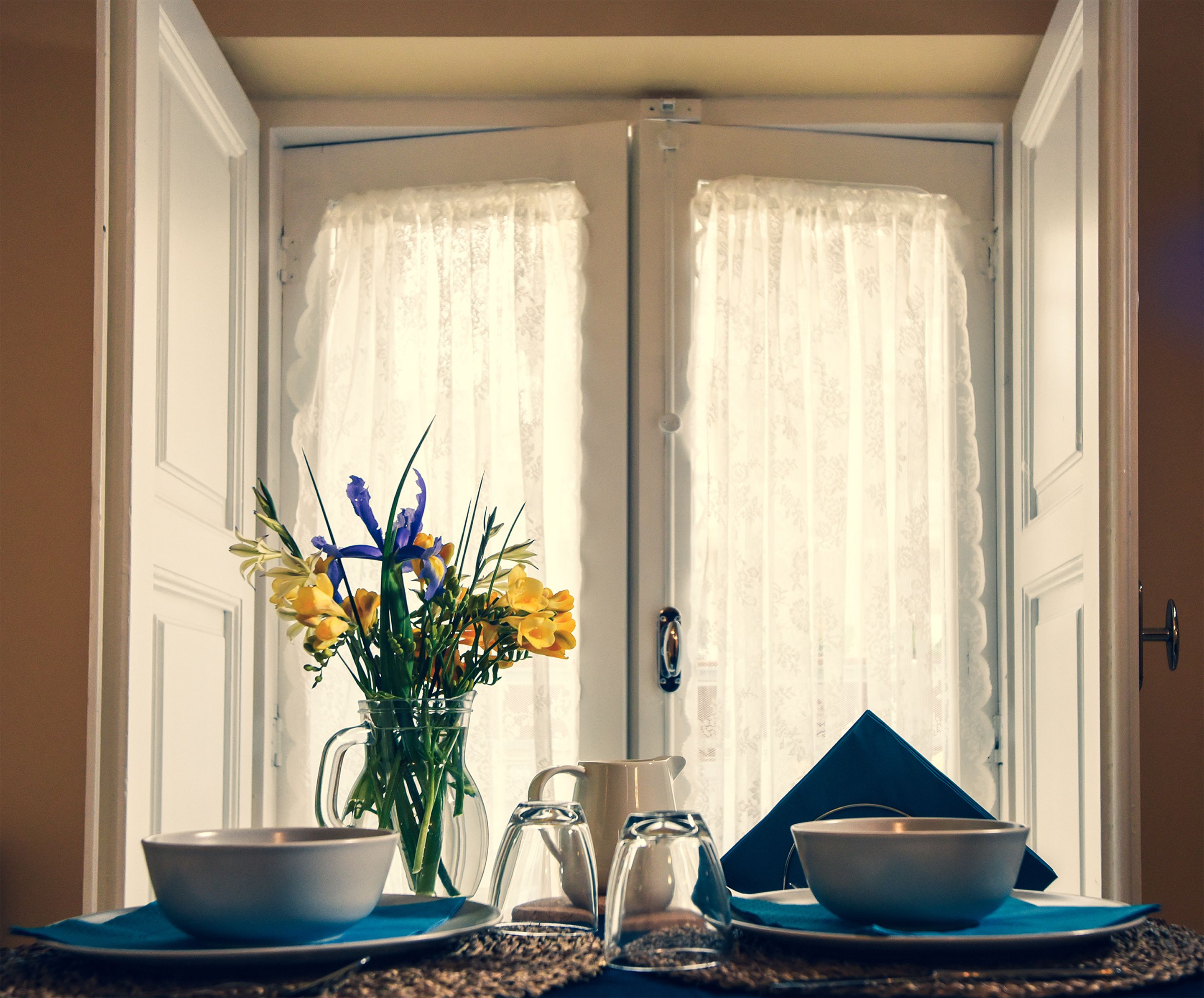 A dining area with a table set for two, a vase of flowers, and a pitcher.