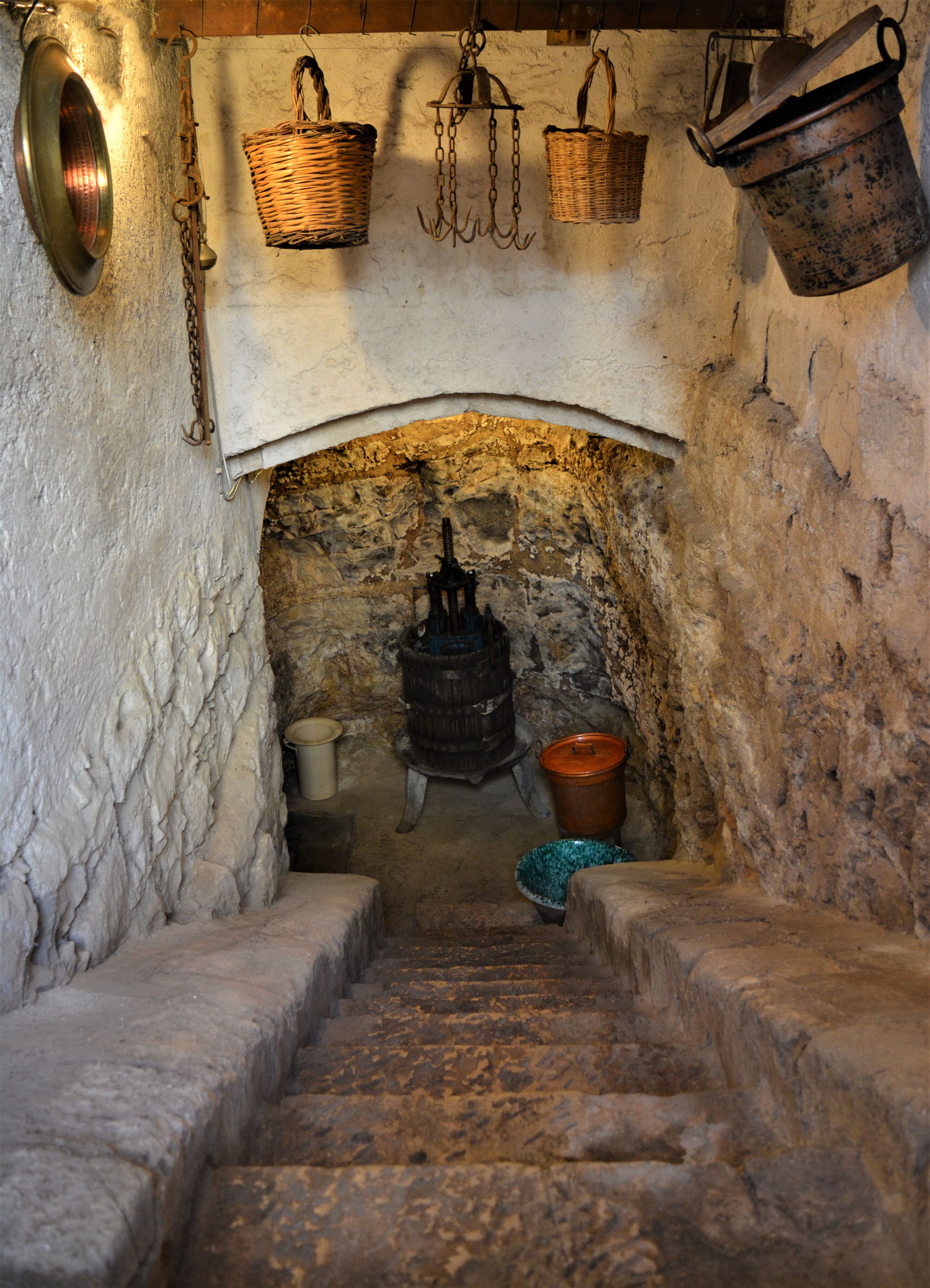 A stone staircase leading to a rustic kitchen with a stone wall, a wooden barrel, and hanging baskets.