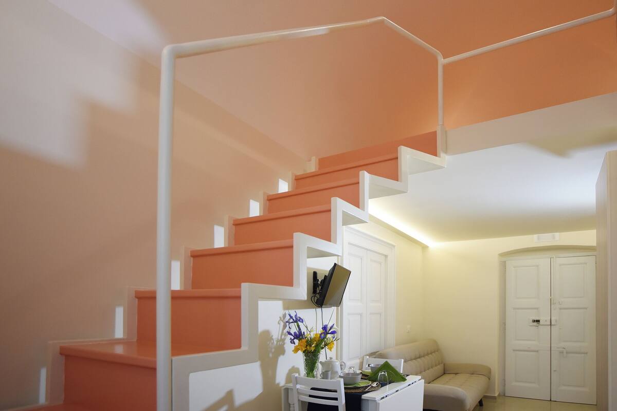Una escalera con barandillas blancas y peldaños anaranjados, una silla blanca, una mesita con un jarrón de flores y un sofá color beige.