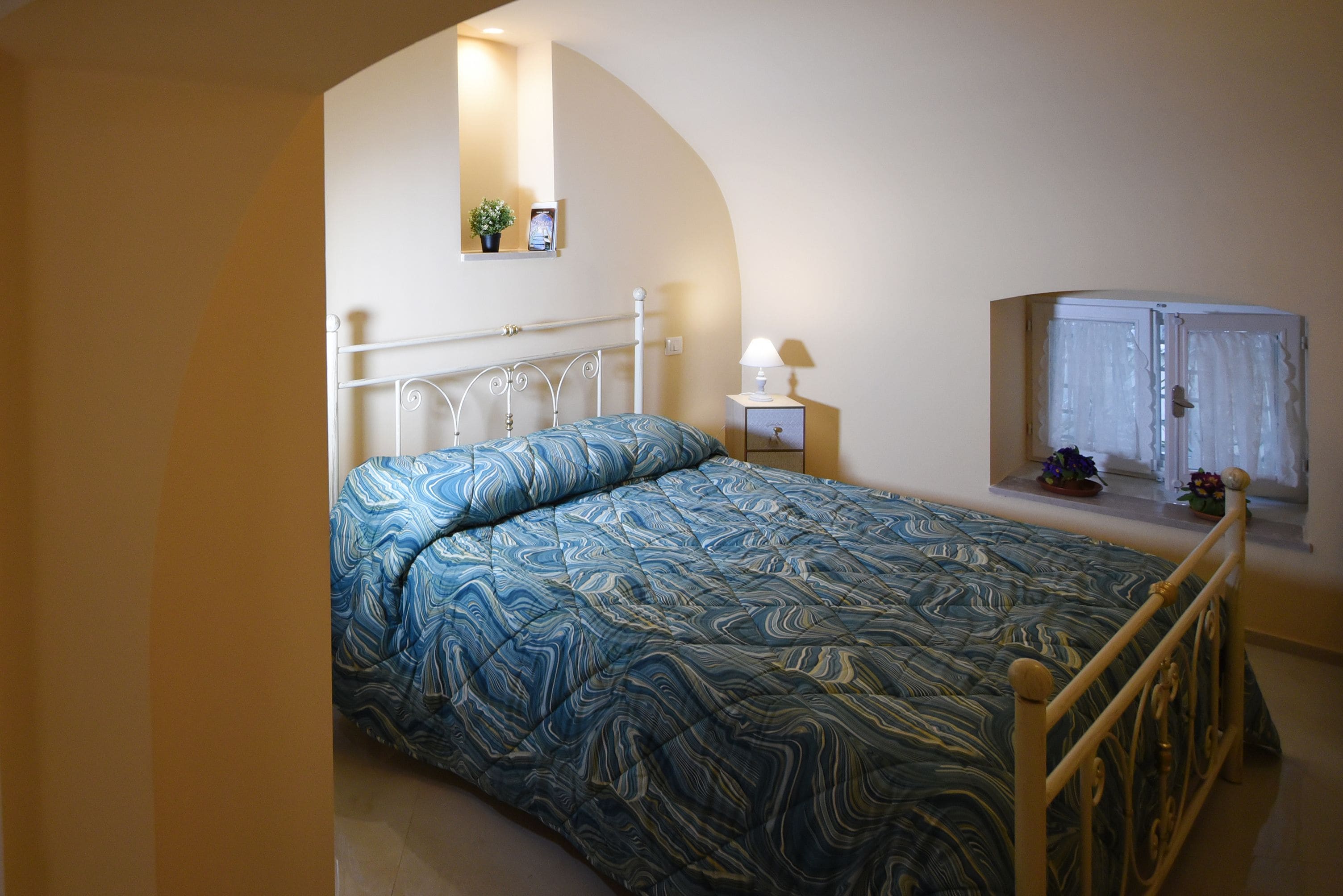 A bedroom with a bed, a nightstand, a lamp, a window with curtains, and a small shelf with a plant.