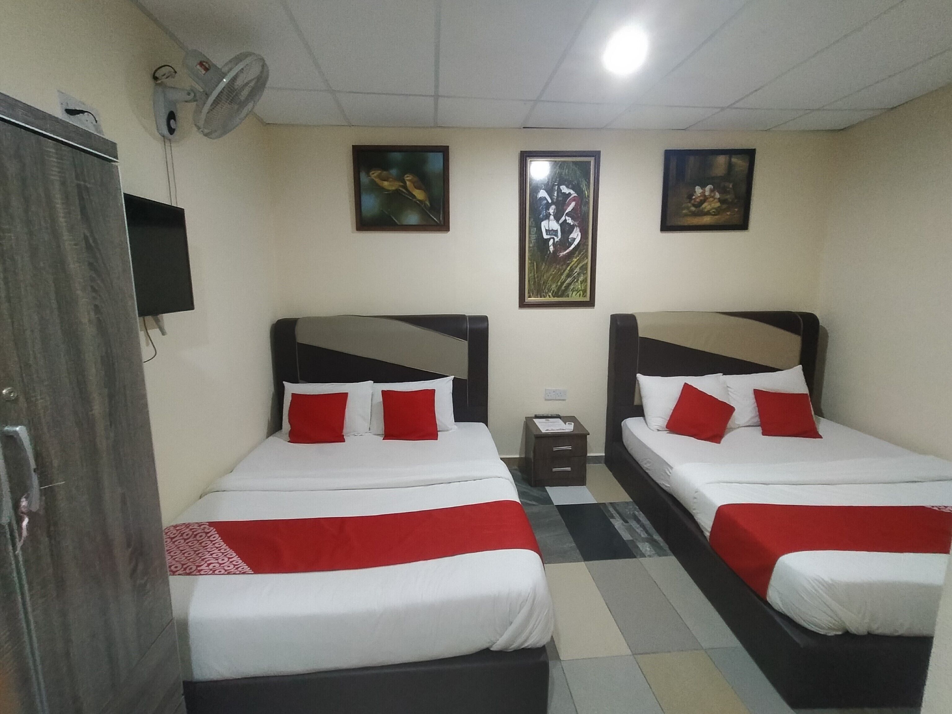 A hotel room with two beds, each with red and white bedding, a small bedside table, a wall-mounted TV, and two framed pictures on the wall.