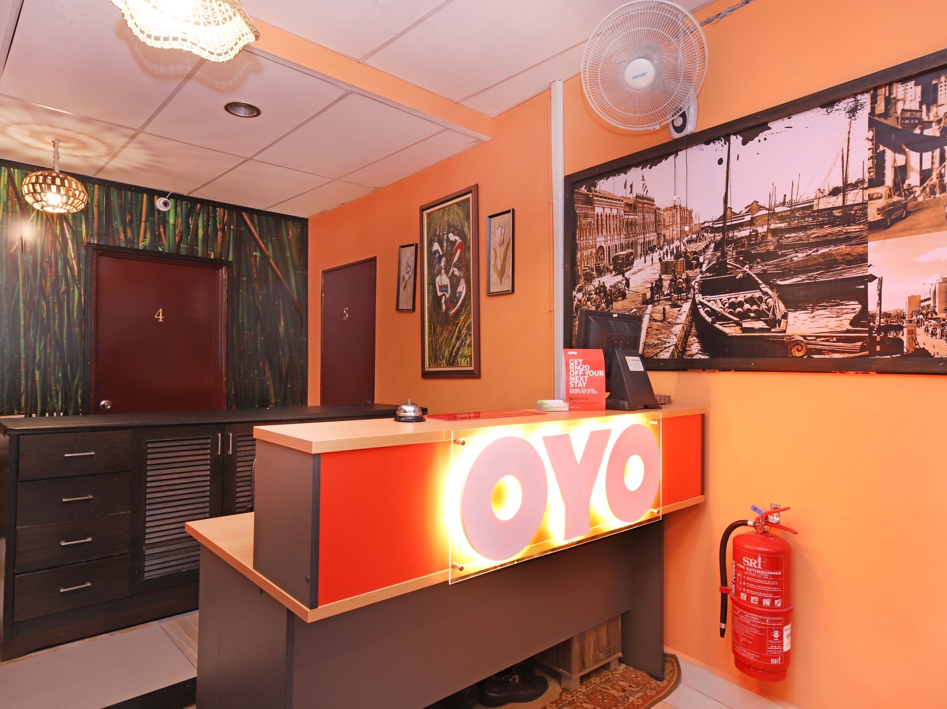 Un mostrador de recepción de hotel con un letrero destacado que dice 'Oya', un extintor y una fotografía grande enmarcada en la pared.