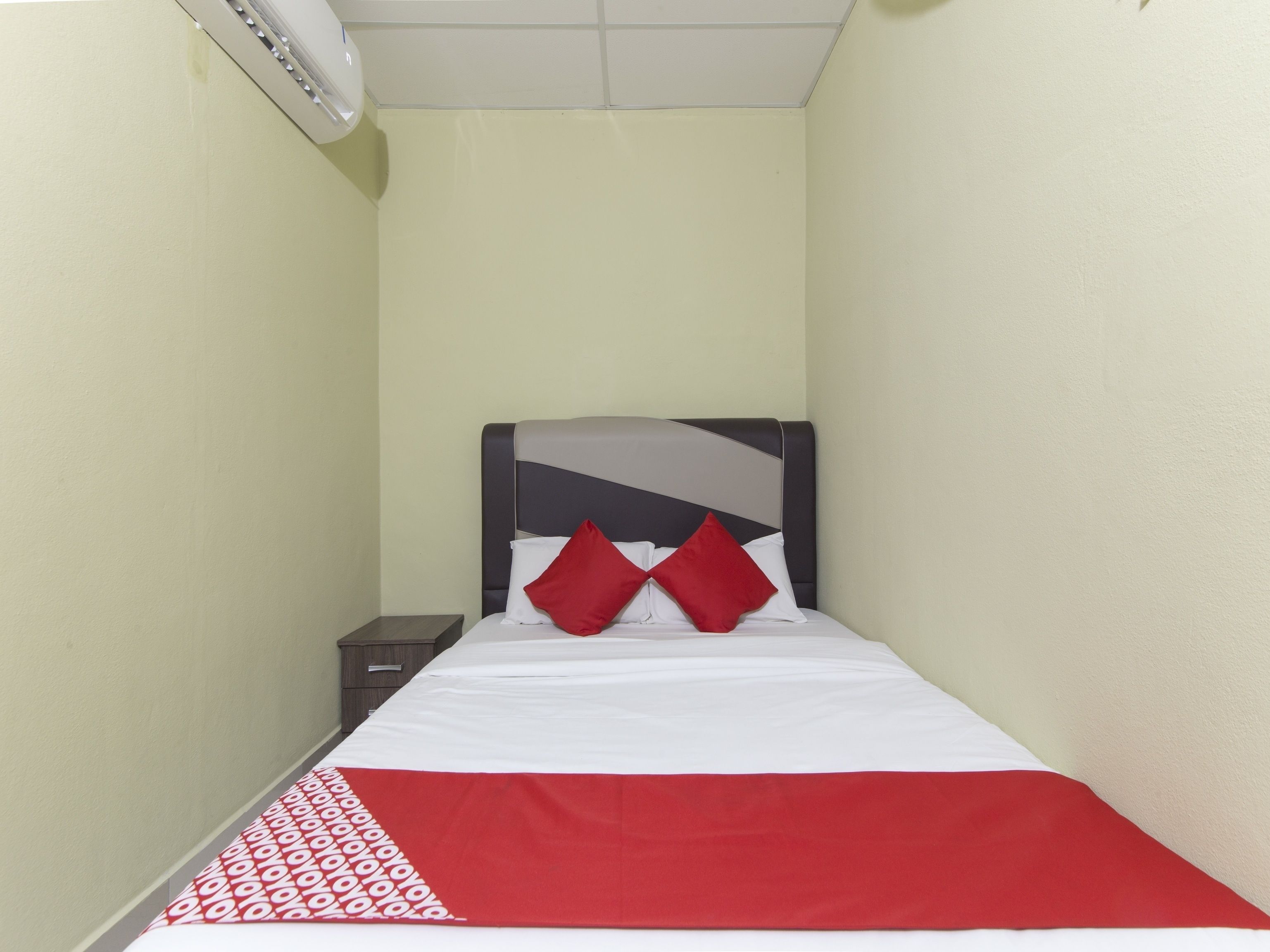 A narrow hotel room with a bed, a nightstand, and a wall-mounted air conditioning unit.