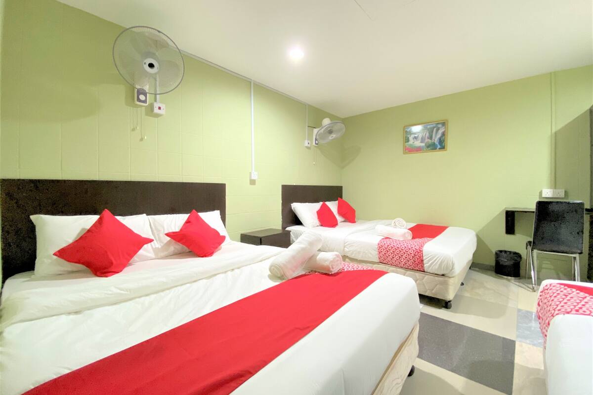 A hotel room with two beds, each with red and white bedding, and a single bed with a red and white blanket. There are two wall-mounted fans and a small table with a chair.