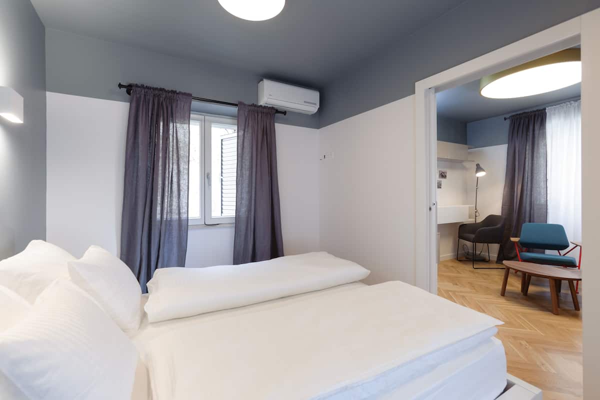 A hotel room with a bed, a desk, a chair, and an air conditioning unit.