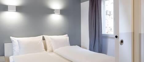 A neatly made bed with white linens, a window with a curtain, and two wall-mounted lights.