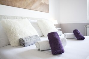 A neatly made bed with white and grey pillows and rolled towels in white and purple. - Apartment Harizma (Belgrade)