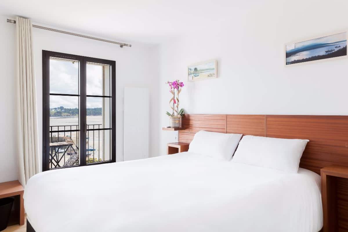 A hotel room with a large bed, a wooden headboard, a small table, a chair, and a window with a view of the outdoors.