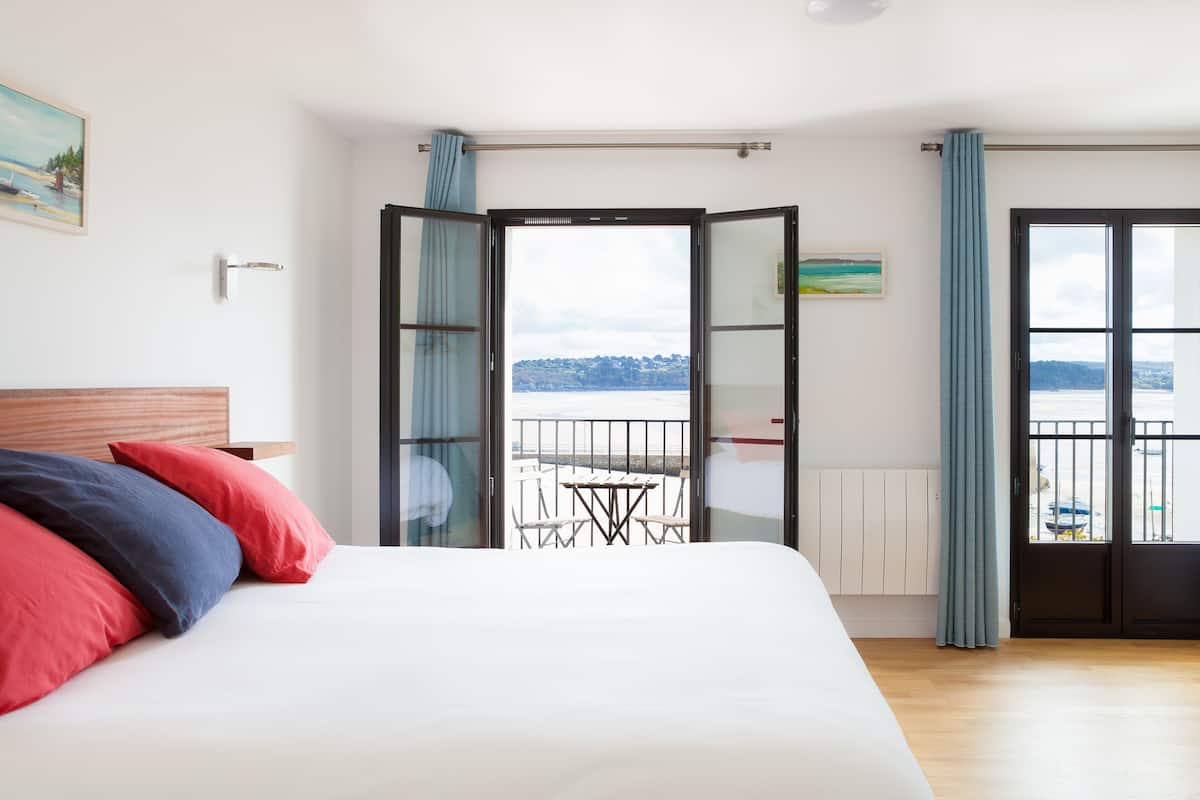 A hotel room with a bed, a view of a balcony with a table and chairs, and a painting on the wall.