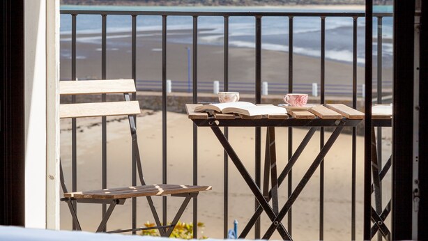 En balkon med udsigt til stranden, et bord med bøger og kopper, samt en seng med en åben bog.