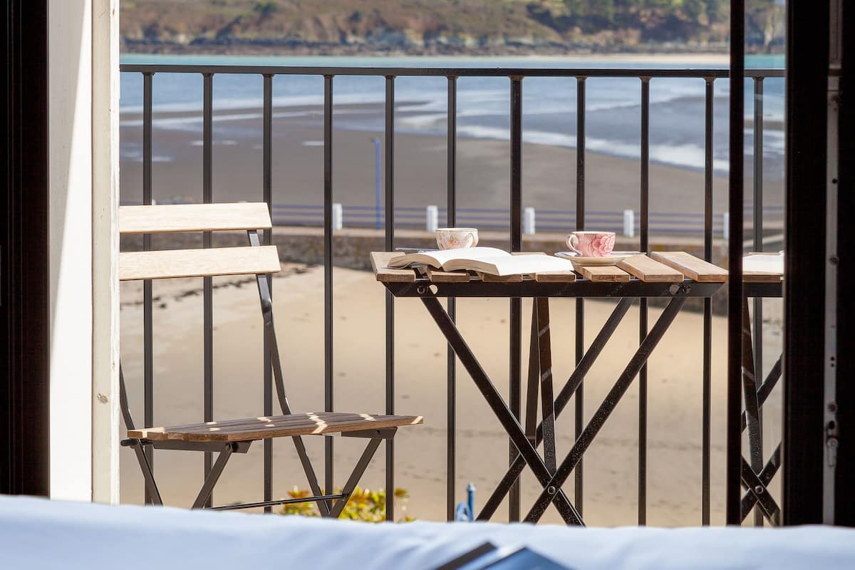 A balcony with a view of the beach, a table with books and cups, and a bed with an open book.