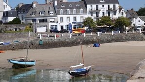 Et kystlandskab med to både fortøjet i rolige farvande, en sandstrand og en række hvide bygninger med balkoner i baggrunden.