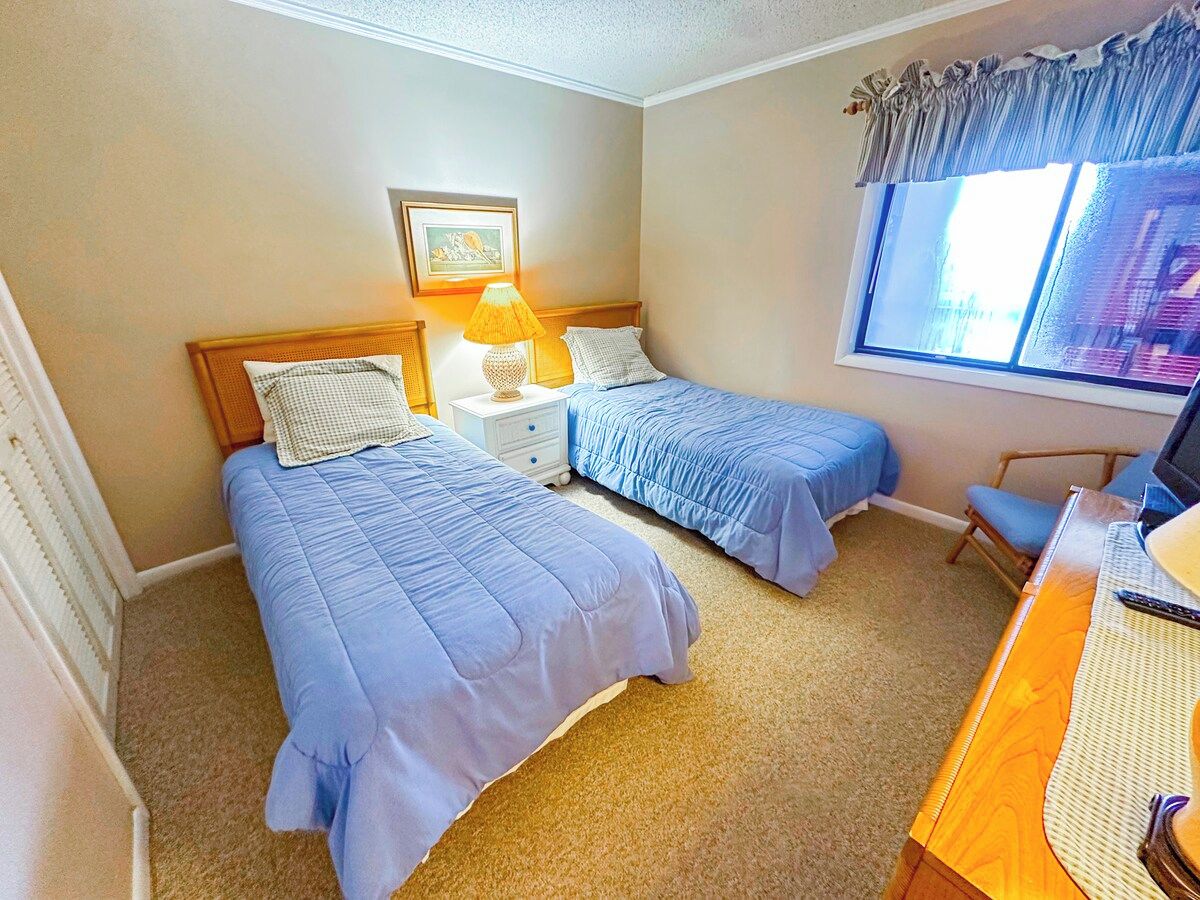 A bedroom with two beds, a desk, a chair, a lamp, and a window with curtains.