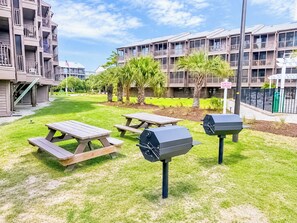 A courtyard with picnic tables and barbecue grills, surrounded by multi-story buildings with balconies. - Experience Tilghman Beach and Racquet Club Unit 207! Oceanfront, Pools, Tennis, Close to Main Street! (North Myrtle Beach)