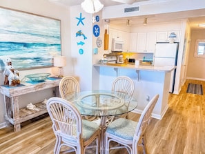 A dining area with a glass table and wicker chairs, a kitchen with white cabinets, and a view of the ocean through a large window.