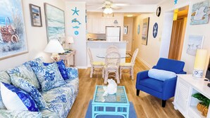 A living room with a blue sofa, a blue armchair, a small table, and a kitchen in the background. - Experience Tilghman Beach and Racquet Club Unit 207! Oceanfront, Pools, Tennis, Close to Main Street! (North Myrtle Beach)