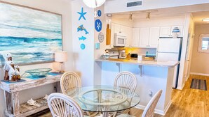 A dining area with a glass table and wicker chairs, a kitchen with white cabinets, and a painting of the ocean on the wall. - Experience Tilghman Beach and Racquet Club Unit 207! Oceanfront, Pools, Tennis, Close to Main Street! (North Myrtle Beach)