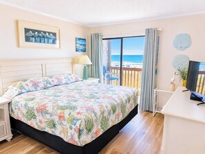 A bedroom with a bed, a desk, a chair, a TV, and a view of the ocean.
