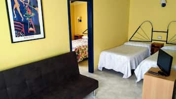 Una habitació amb un sofà marró fosc, dos llits individuals amb roba de llit blanca, una tauleta de nit de fusta amb un televisor, i una obra d'art emmarcada a la paret.