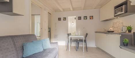 A modern living room with a grey sofa, a dining table with chairs, a kitchen area with a microwave, and a ceiling with exposed beams.