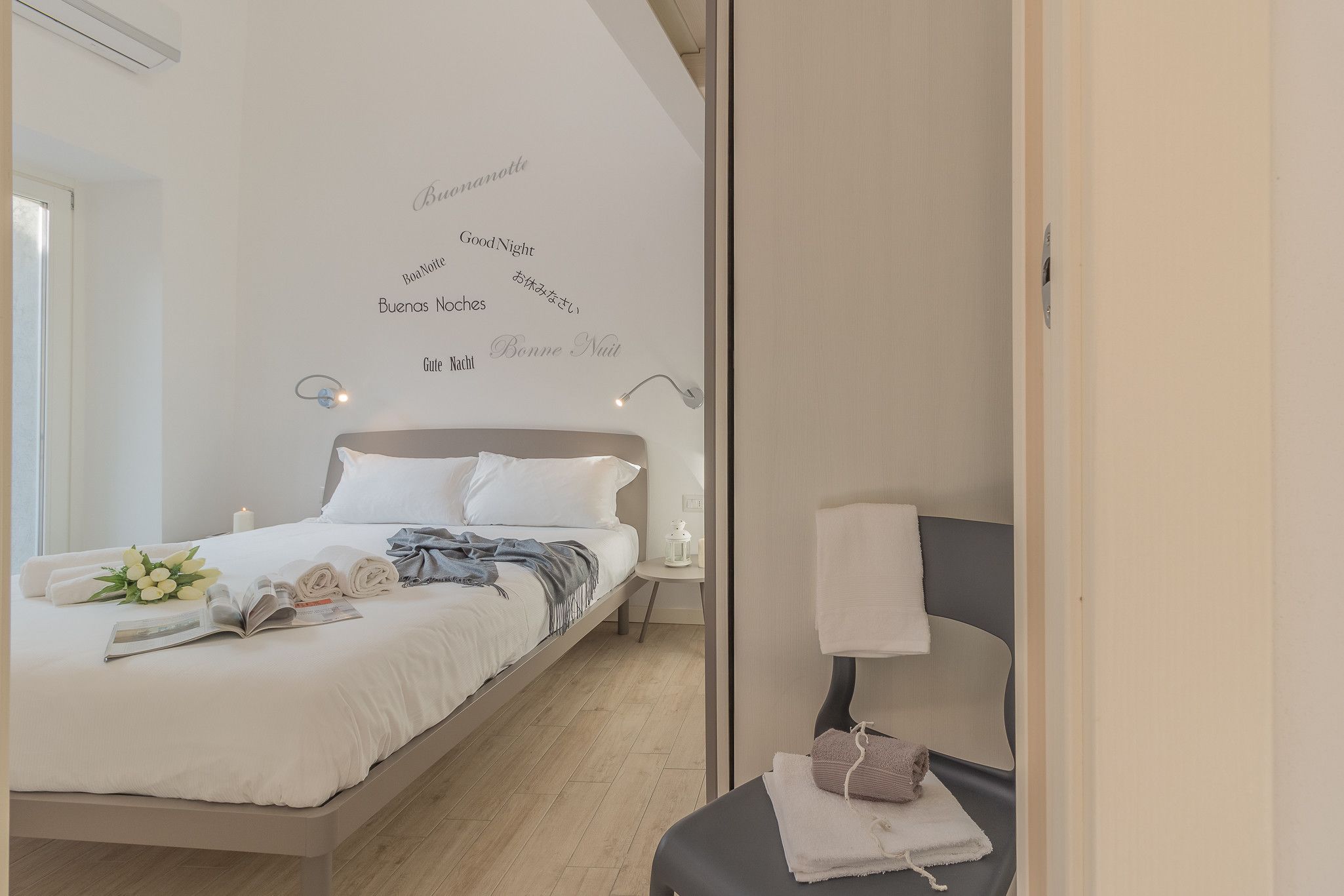 Una cama bien hecha con sábanas blancas y un ramo de flores, una mesita de noche con una lámpara, y una pared con mensajes de 'Good Night' en varios idiomas.