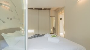A neatly made bed with white linens, a grey jacket, and a rolled towel. - Pretty Home (Varenna)