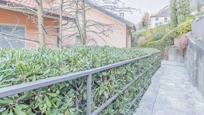 A narrow pathway flanked by a metal railing and dense green foliage, leading to a building with a red-tiled roof and a stucco exterior. - The Apple House (Varenna)