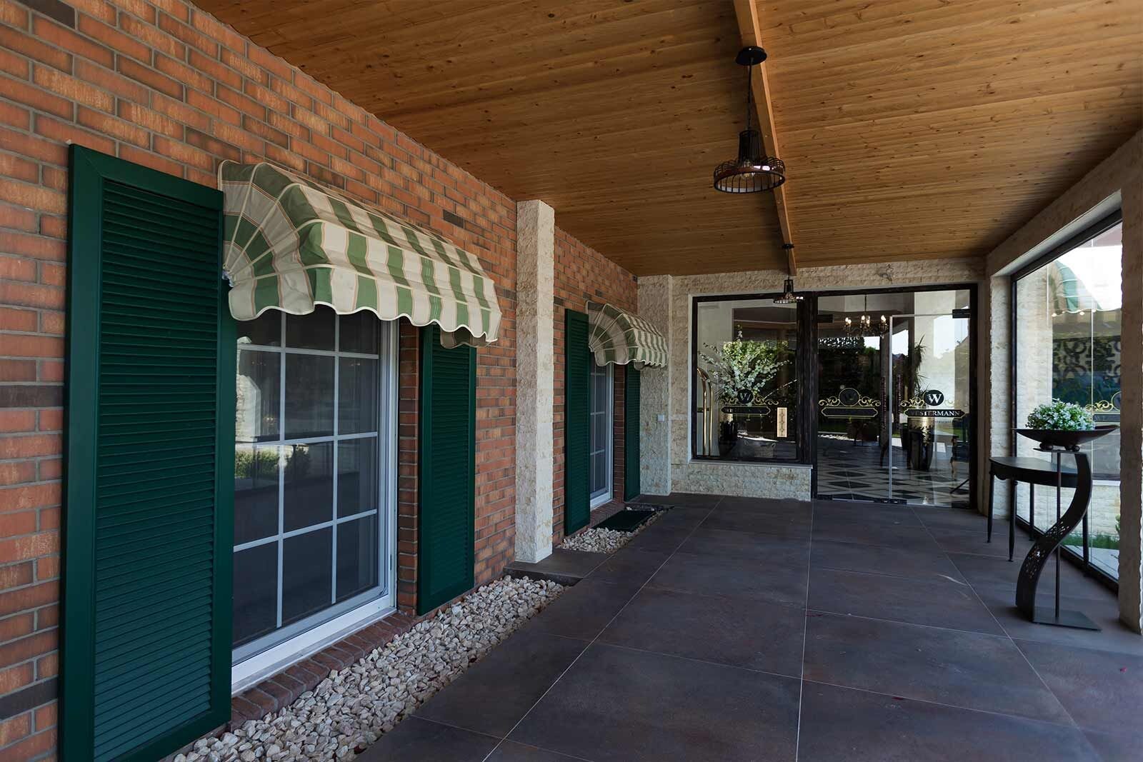 A covered outdoor area with brick walls, green shutters, and a tiled floor leading to a glass door entrance.