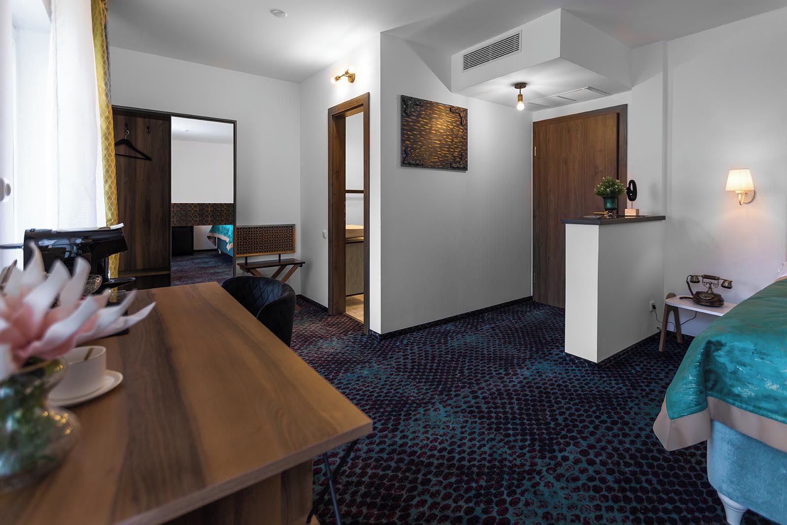Een hotelkamer met een houten bureau, een bed met een blauwgroene spreidek, een kleine tafel met een lamp, een kapstok en een kunstwerk aan de muur.
