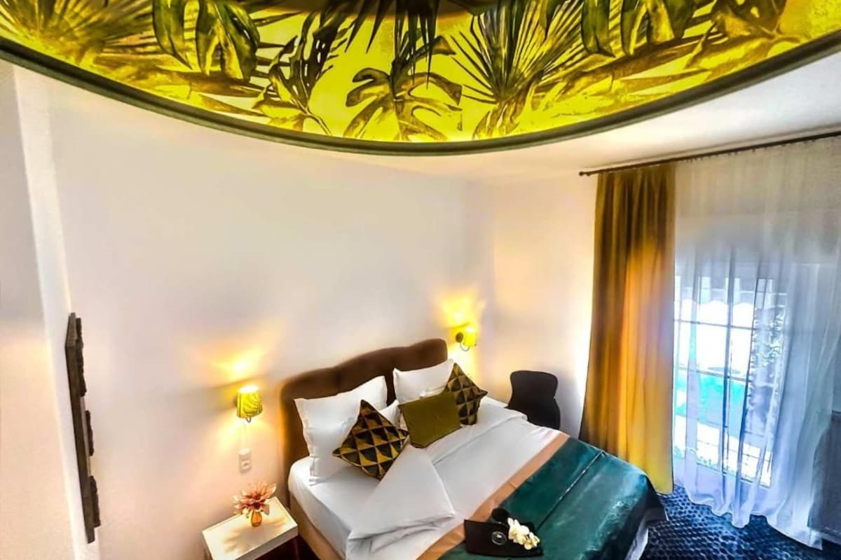 Una habitación de hotel con cama, lámparas de noche, mesita de noche y ventana con cortinas.