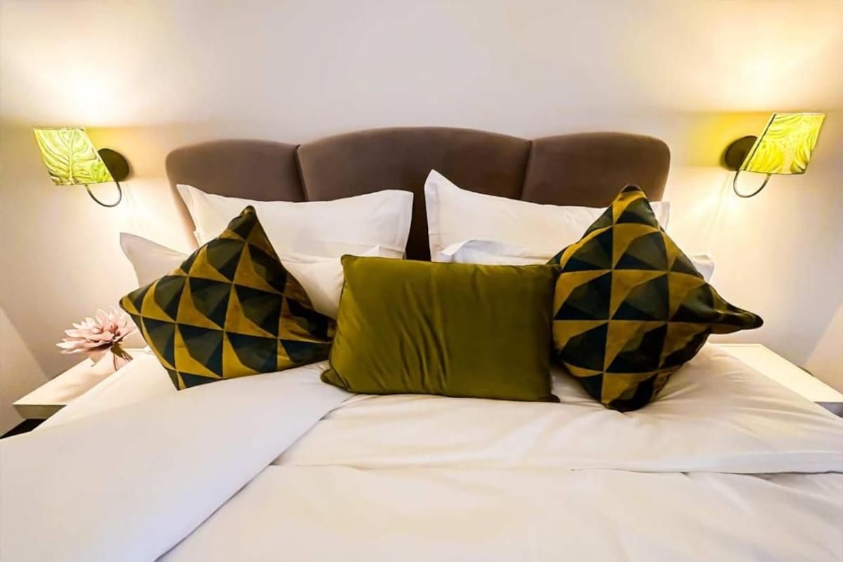 Una cama con sábanas blancas, un cabecero tapizado marrón y almohadones decorativos con diseños geométricos.