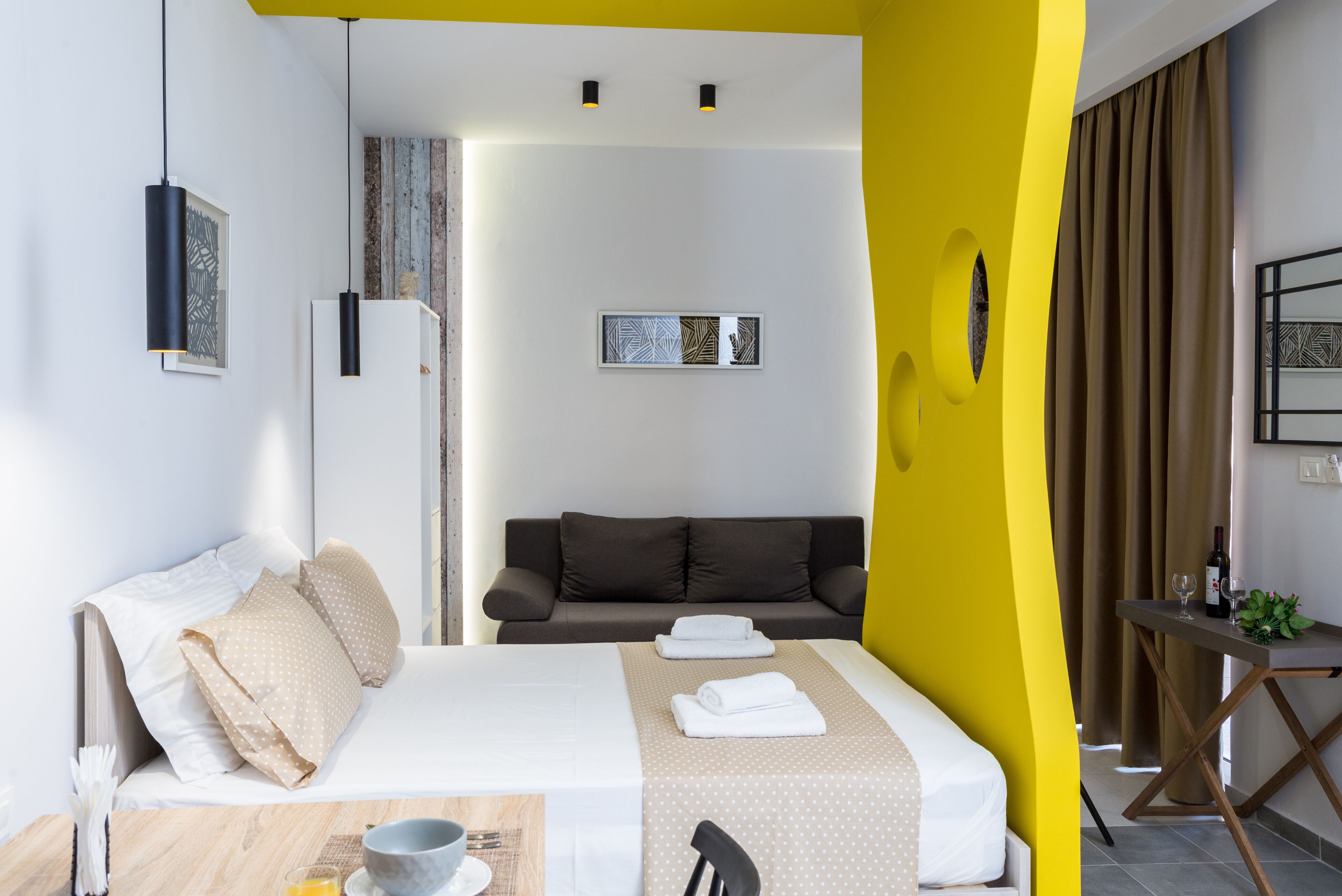 A modern hotel room with a bed, a sofa, a dining table, and a small table with a bottle and glasses.