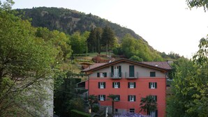 A multi-story building with a red facade, green shutters, and a balcony. It is surrounded by lush greenery and a hillside in the background. - Bellagio Bed & Breakfast (Bellagio)