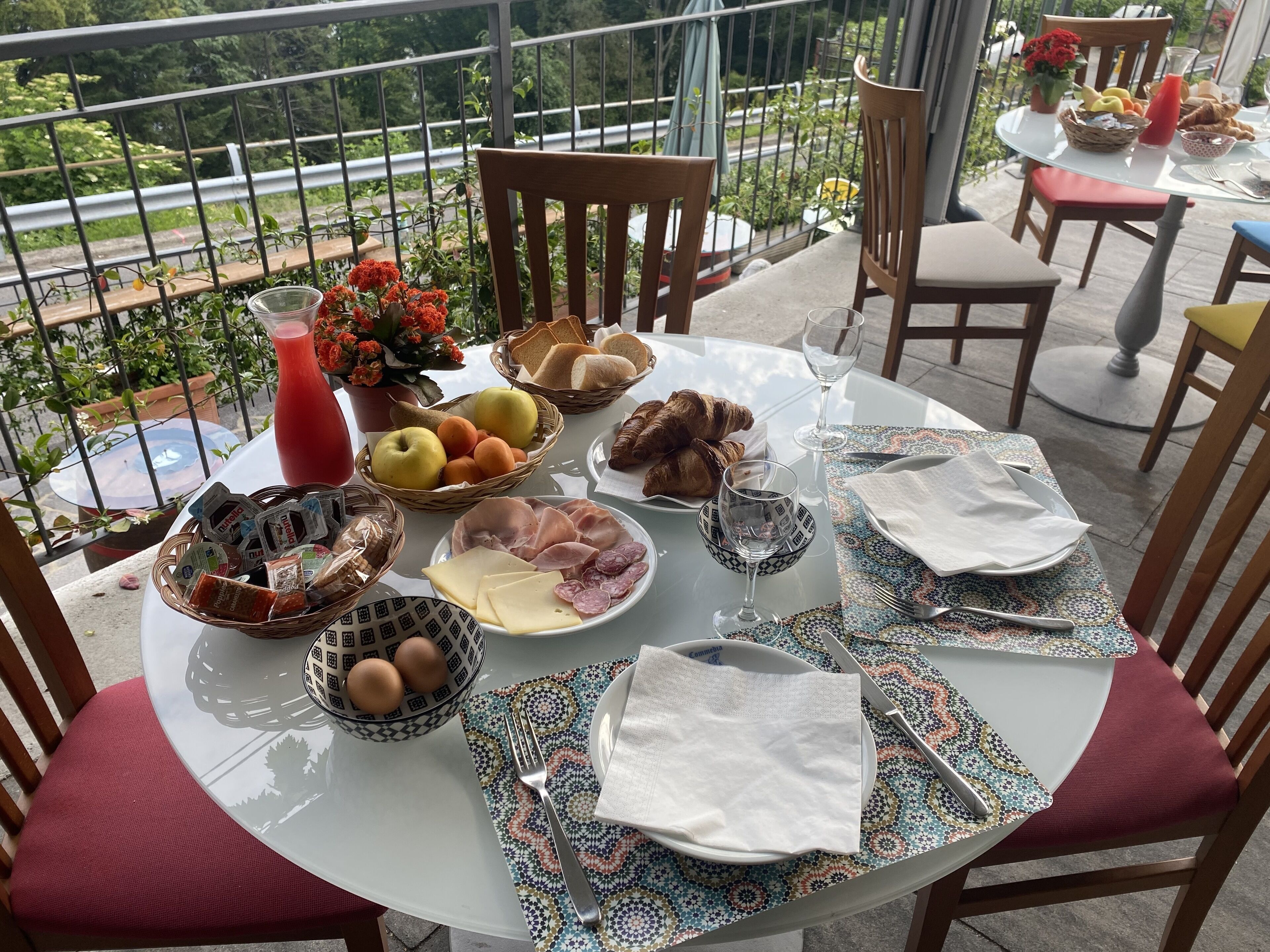 Een ronde tafel gedekt voor het ontbijt met verschillende soorten eten, een glas rode sappen en een uitzicht op groen en een weg op de achtergrond.